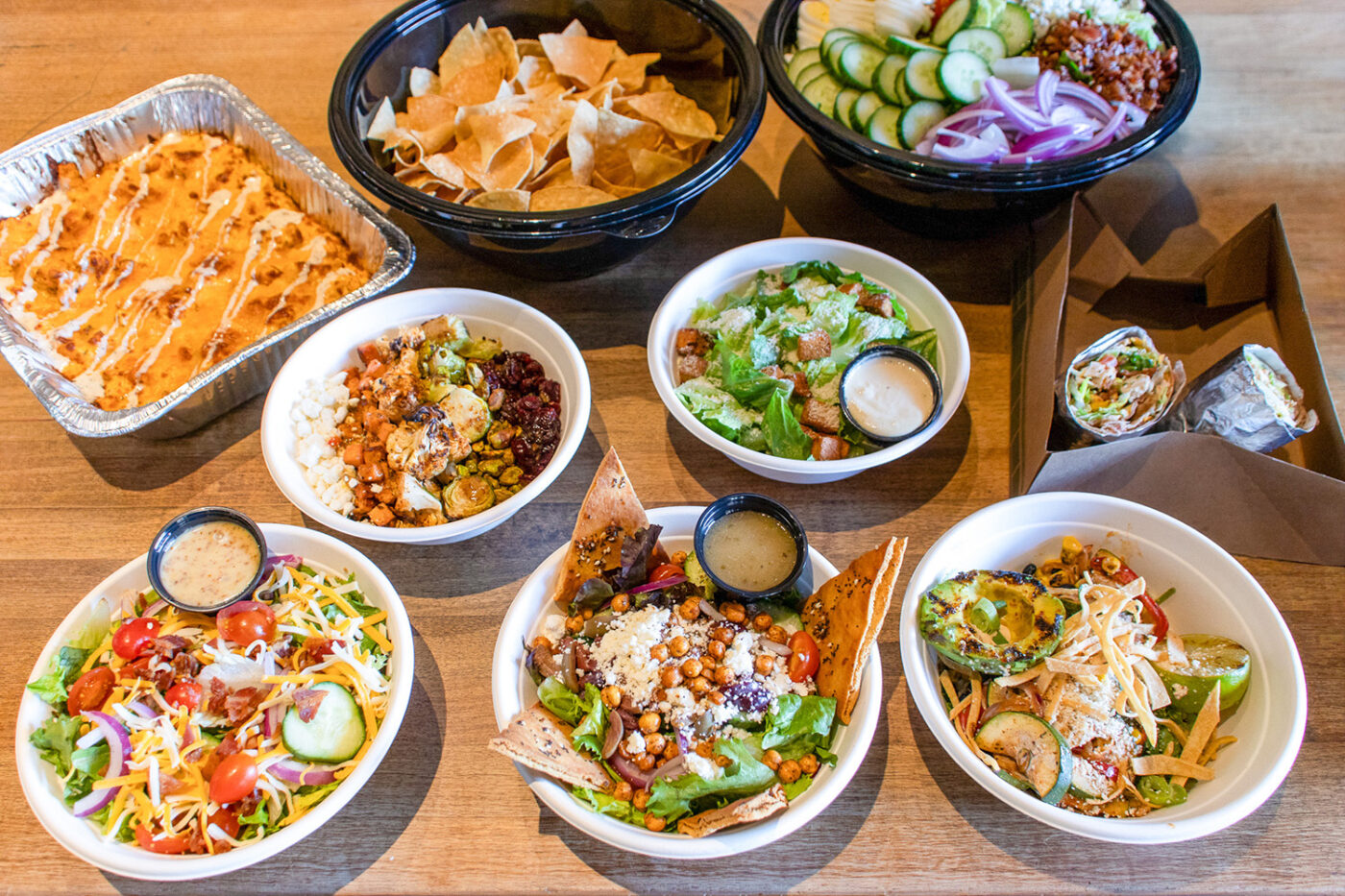 Catering spread featuring Cheesy Mac, House Salad, Greek Salad, Southwest Avocado Bowl, Roasted Vegetable Bowl, Chicken Caesar Wrap and a side of House-made Chips