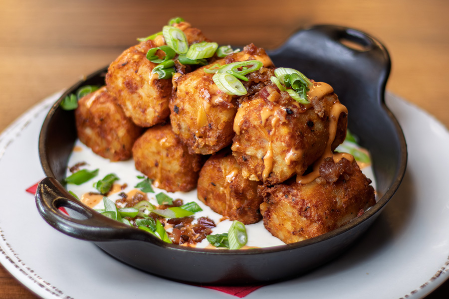 Loaded Jumbo Tater Tots made with applewood-smoked bacon, queso sauce, topped with scallions and a spicy aioli